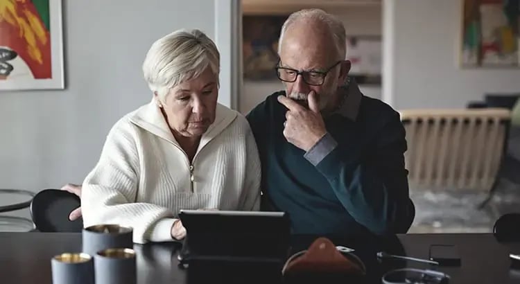 An elderly couple sitting together, looking at a laptop. They appear to be engaged in a focused discussion or review.