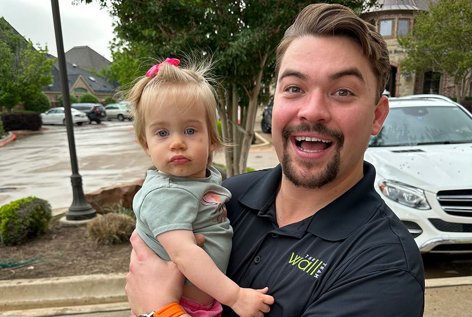 A man in a polo shirt holding a baby girl with a white car in the background