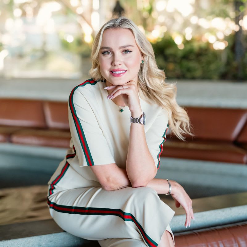 Ilona Laor seated on a bench in a cream outfit with green and red stripes.