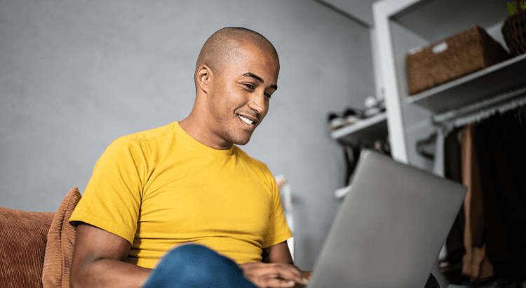 A smiling man wearing a bright yellow t-shirt, sitting and using a laptop.