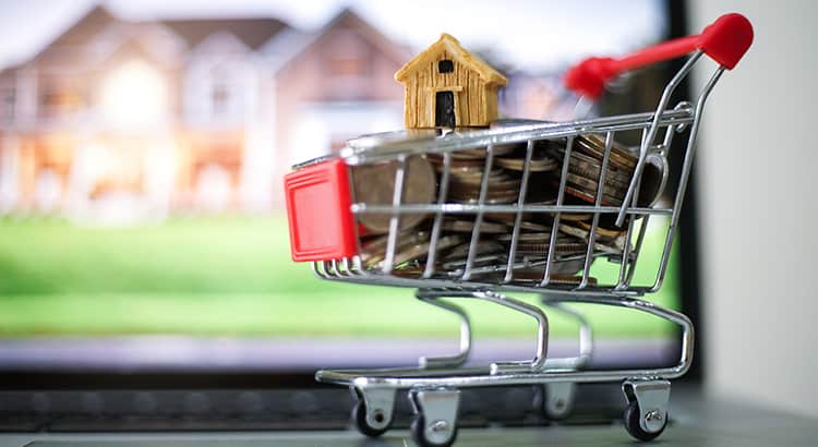  A small shopping cart with a tiny wooden house inside, placed against a blurred green background. This image likely symbolizes purchasing a home.