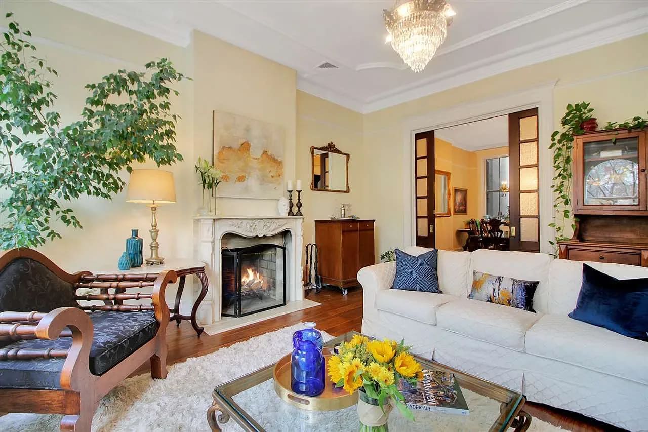 A living room with a fireplace, a painting on the wall, a couch, several houseplants, and a door leading to a dining room.