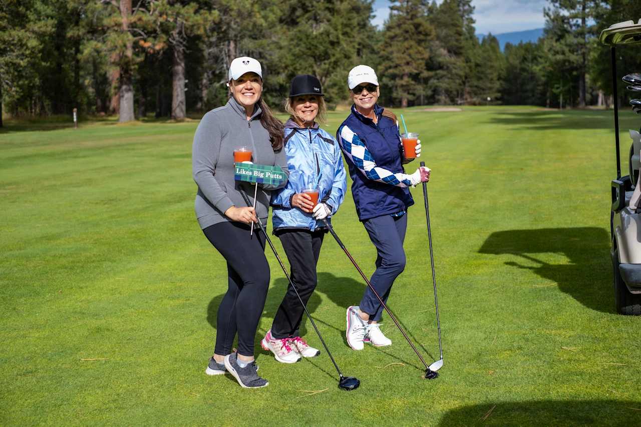 Three women holding golf clubs and drinks on a green golf course.