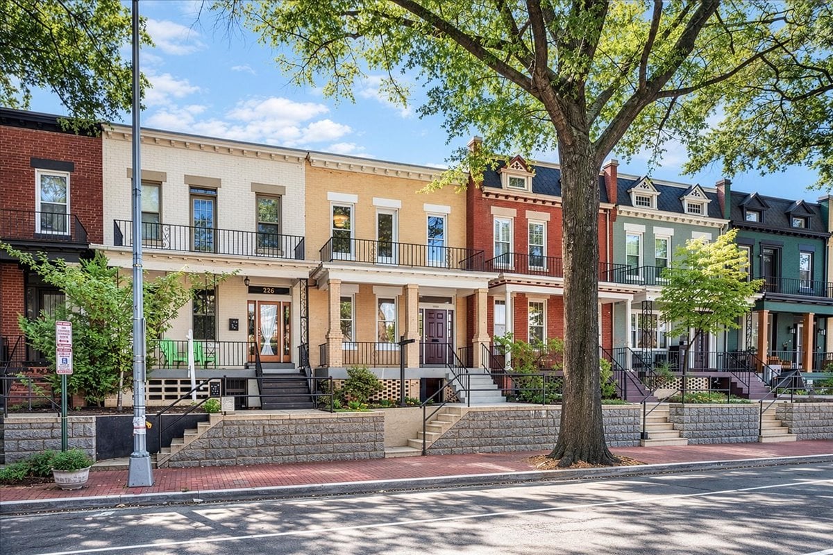 Attached brick townhouses with raised porches and stone retaining walls beneath a large leafy tree.