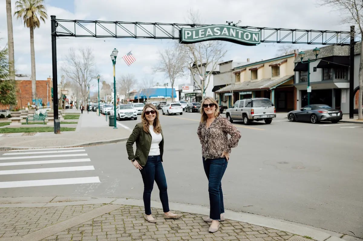 Two women stand beneath the Pleasanton sign on a downtown street.