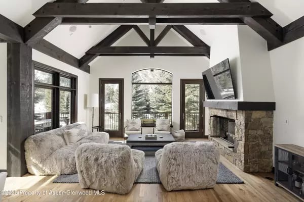 Living room with exposed timber beams, a stone fireplace, and large windows overlooking trees.