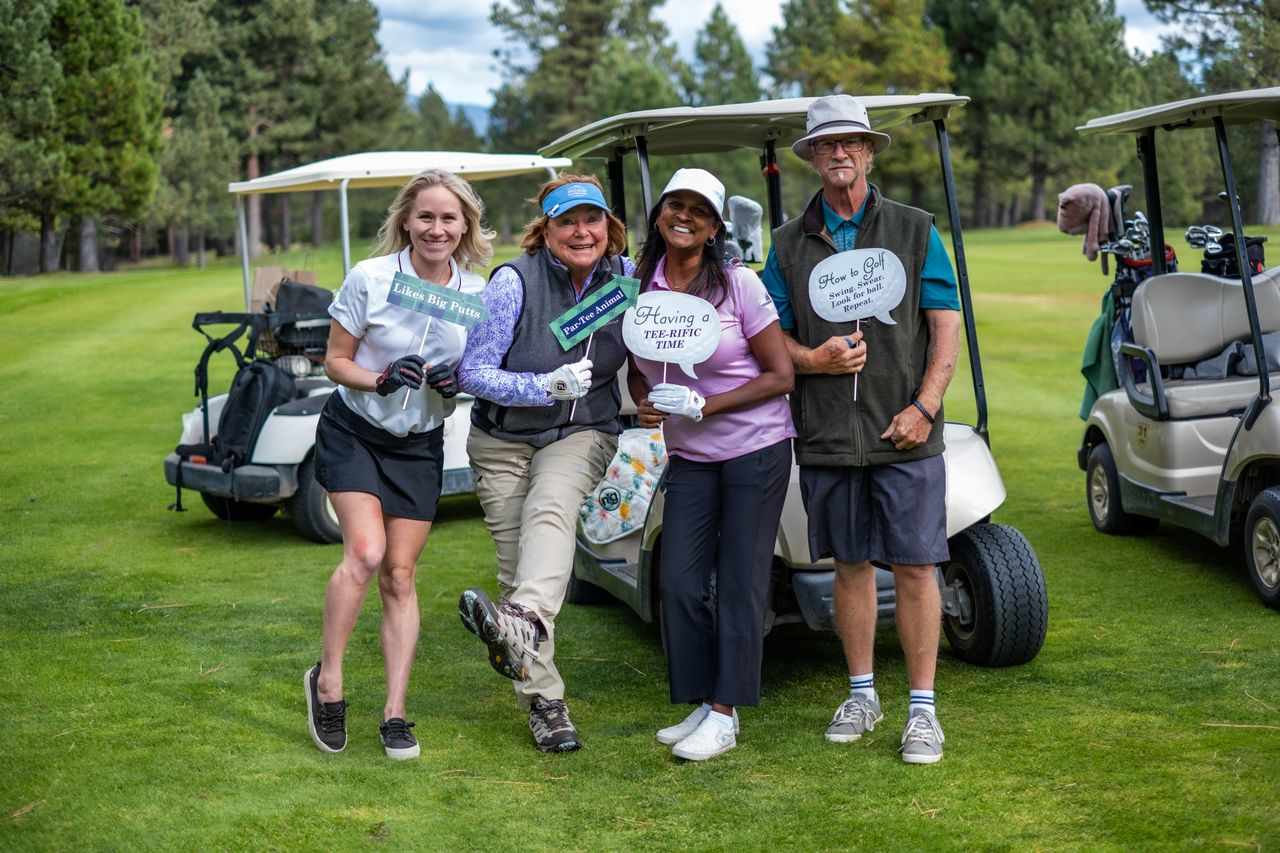 Four adults pose with golf-themed signs beside golf carts on a green course.