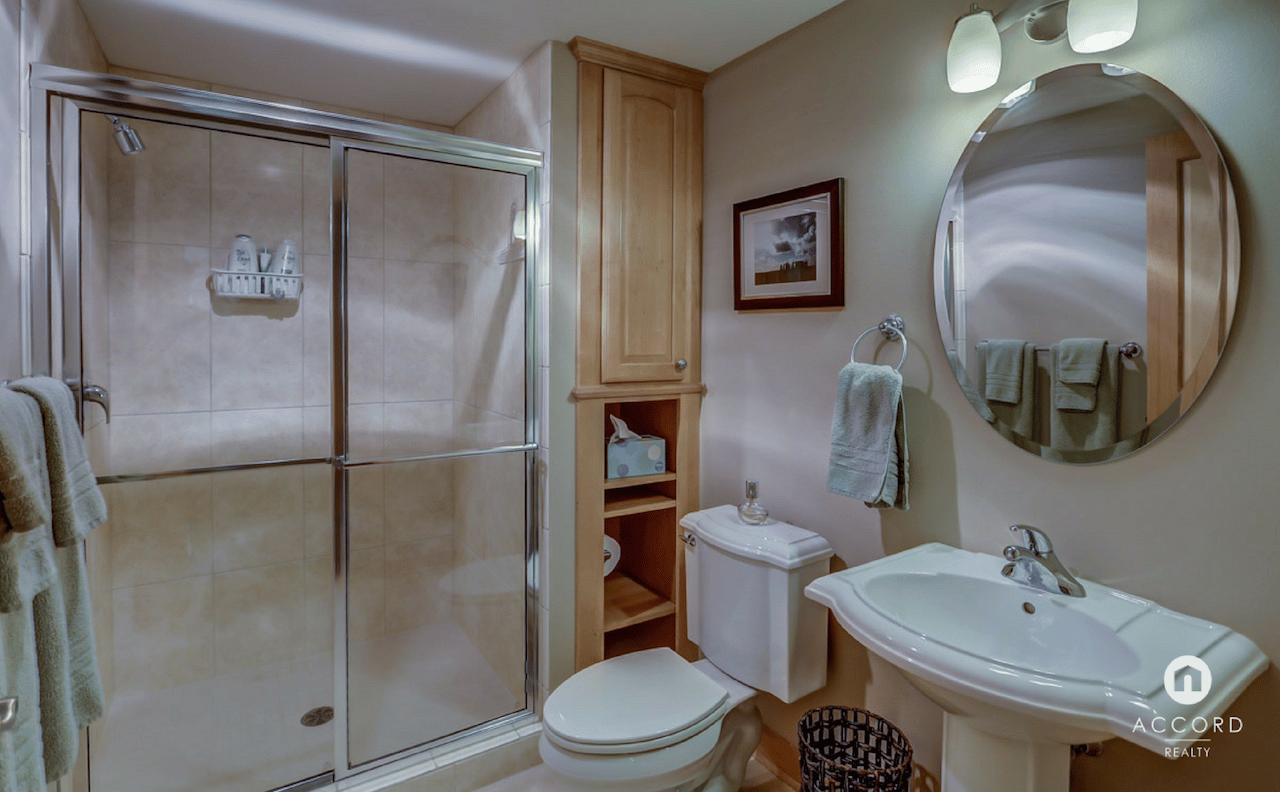 Condo bathroom with a glass shower, white sink, wooden cabinet, and large round mirror.