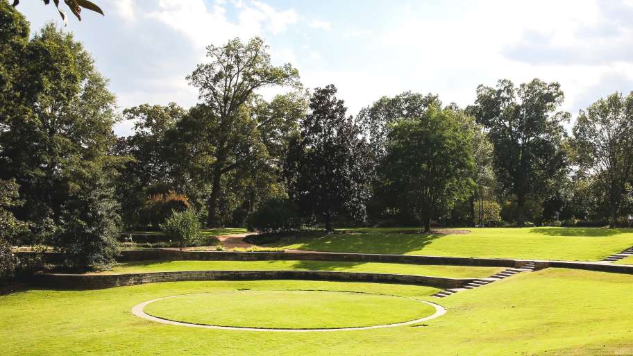 A serene park scene featuring a circular stone structure on lush green grass, surrounded by trees under a partly cloudy sky.