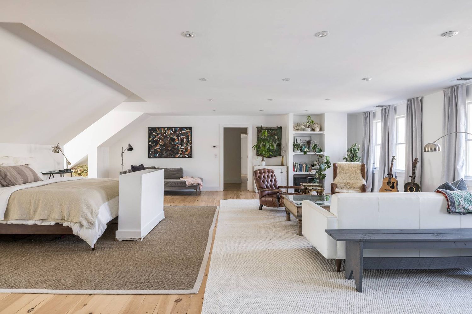 Bright loft bedroom and living area with a sloped ceiling, white sofas, and tall windows.