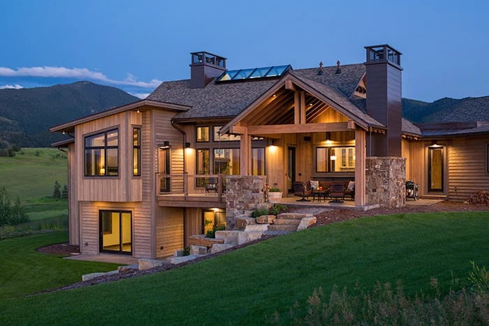 Two-story timber-and-stone home glowing at dusk, with mountain foothills behind it.