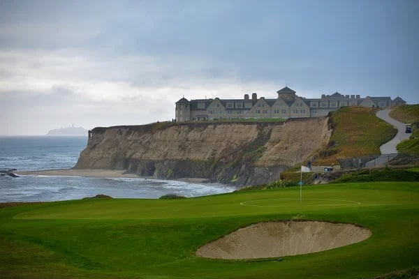 Coastal golf course and bunker below ocean cliffs, with a resort building atop the bluff.