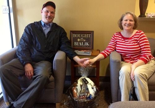 A smiling couple seated in armchairs, holding hands beside a Bialik Real Estate sign.