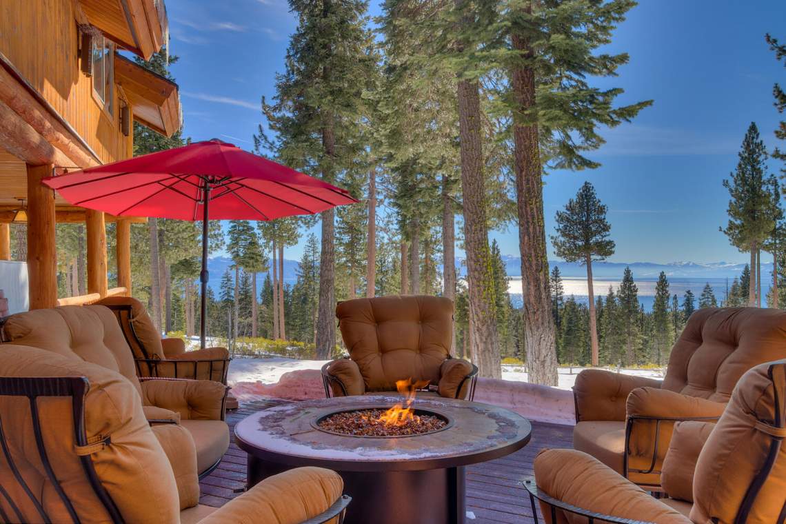 Furnished patio with a circular fire pit, red umbrella, and view of Lake Tahoe through snowy pines.