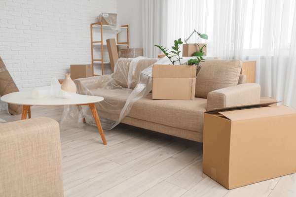 Bright living room with cardboard moving boxes and furniture wrapped in protective plastic.