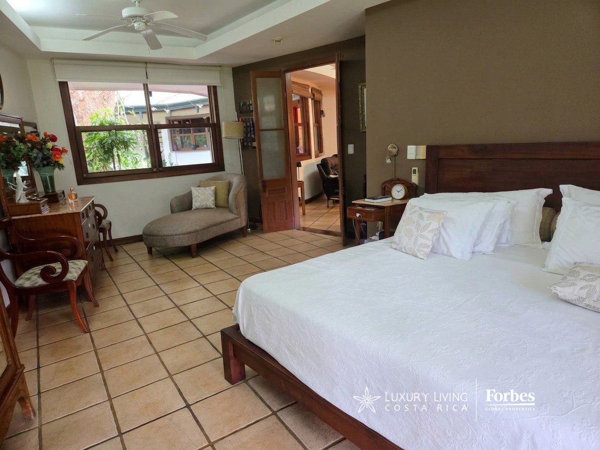 Furnished villa bedroom with a large white bed, dark wood furniture, and a window overlooking greenery.