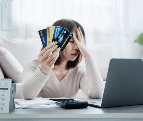 Woman holds credit cards at a desk beside a laptop, pressing her forehead with her other hand.