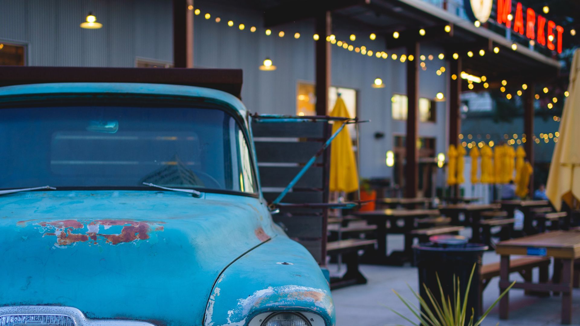 Weathered turquoise pickup parked beside a restaurant patio lit by string lights.