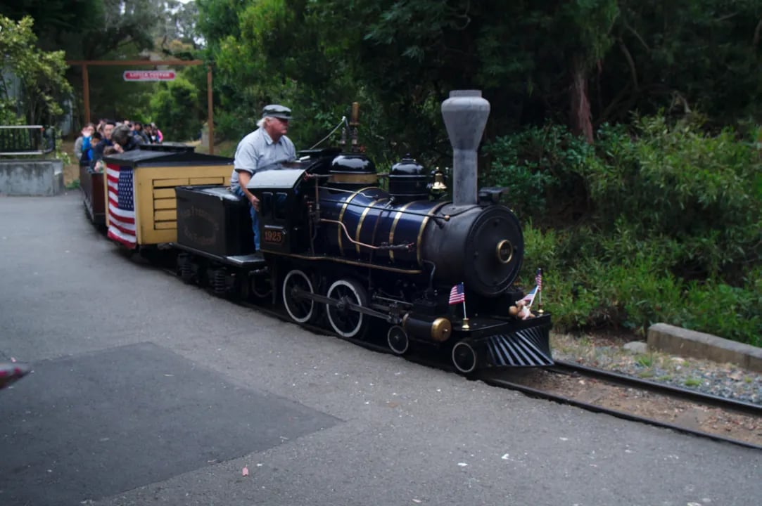 Children and families riding the historic Little Puffer steam train with the zoo's lush landscaping and animal exhibits visible in the background.