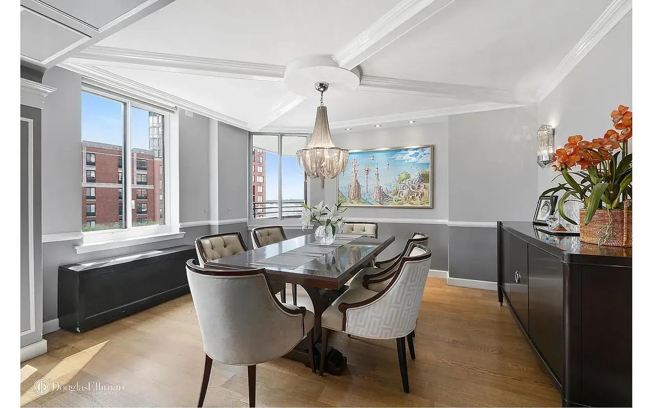 Dining room with a crystal chandelier, long table, upholstered chairs, and large windows.
