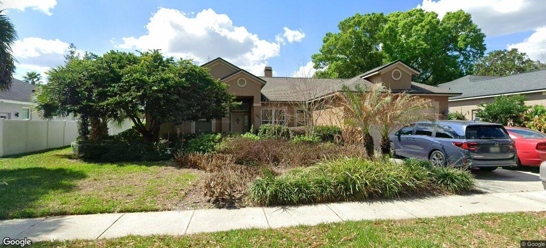Single-story brown house with gabled rooflines, mature trees, and cars parked in the driveway.