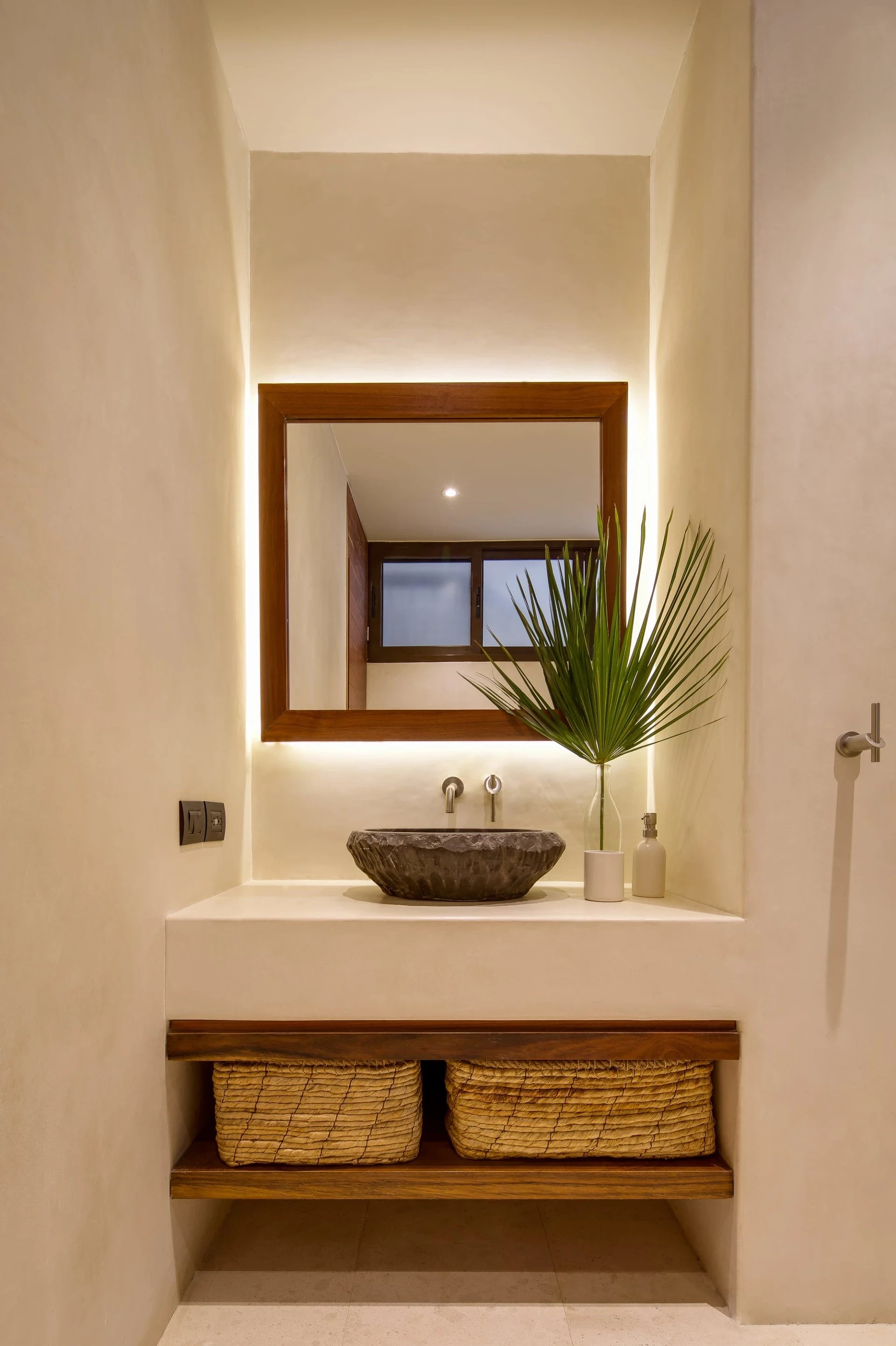 Bathroom vanity with a stone vessel sink, illuminated wood-framed mirror, palm frond, and woven baskets.