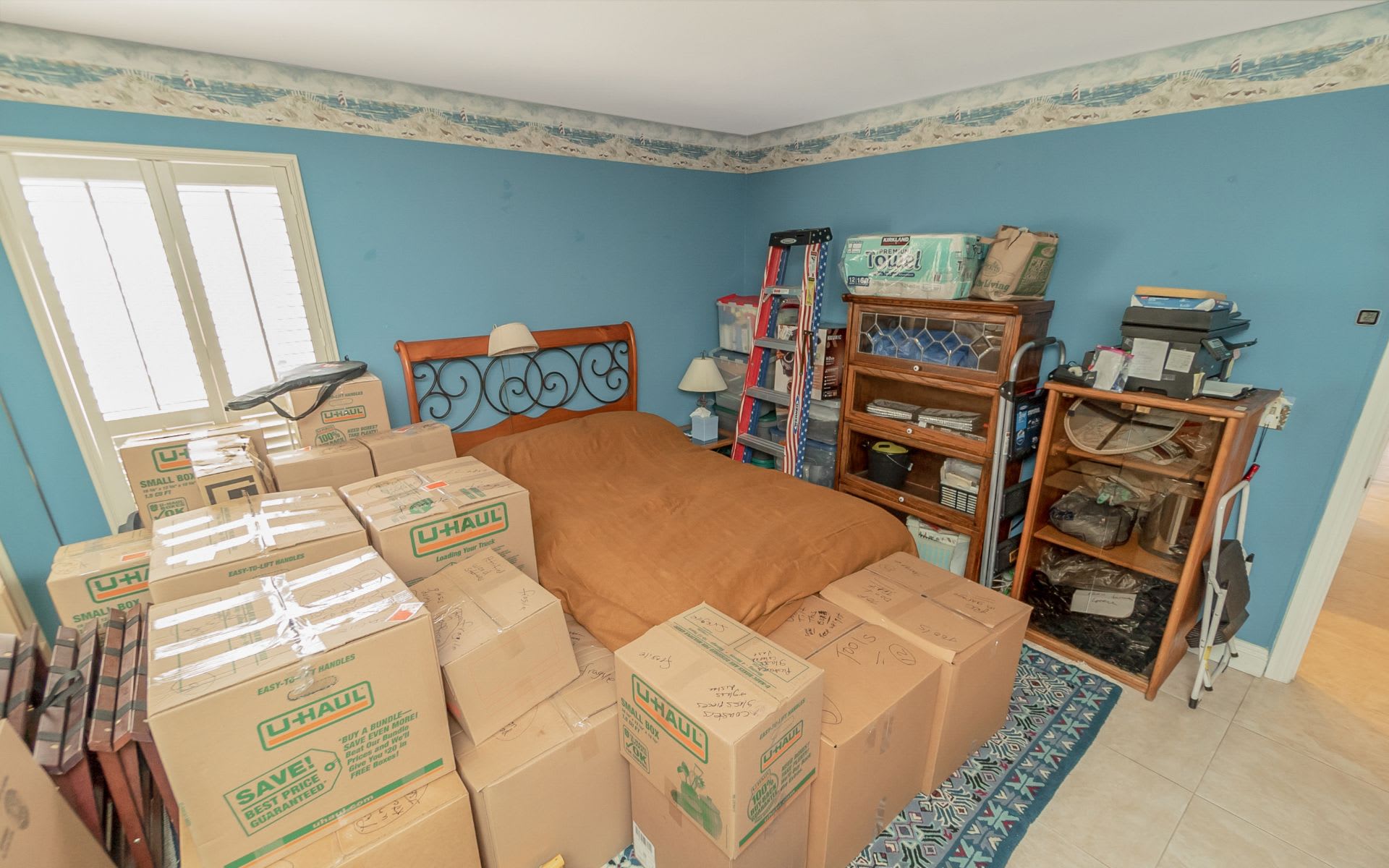 Bedroom crowded with moving boxes around a bed, with blue walls and wooden cabinets.