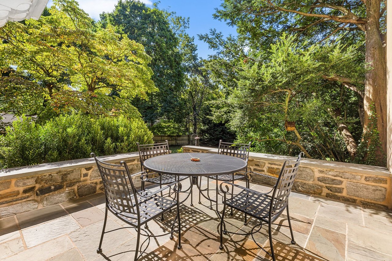 Stone patio with a round outdoor dining set, low stone wall, and mature trees.
