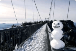 Snowy suspension bridge with a snowman beside the railing and mountains in the distance.