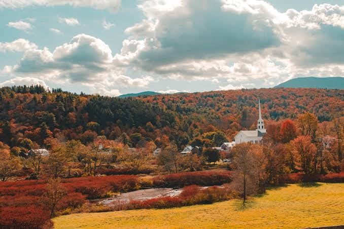 Autumn countryside with a white-steepled church, colorful wooded hills, stream, and golden field.