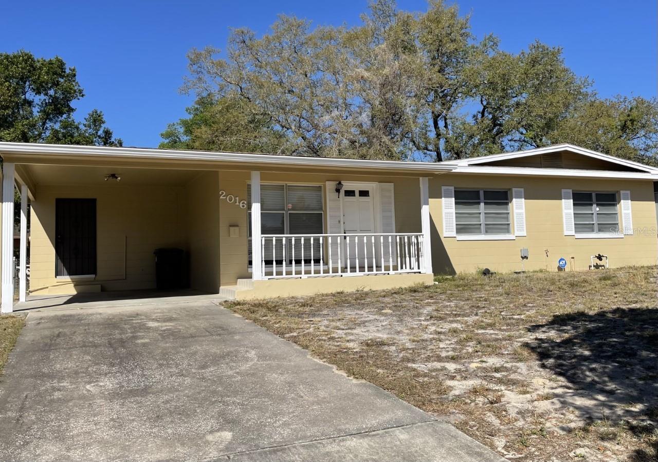 Single-story yellow house with a covered carport, white porch railing and concrete driveway.