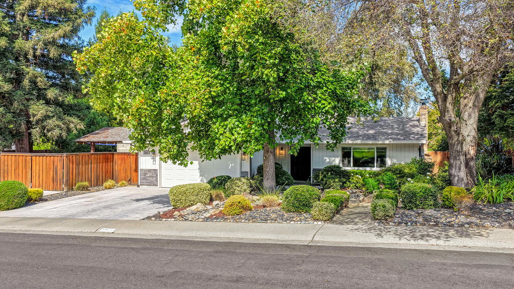 Single-story white home with a paved driveway, wooden fence, and mature trees.