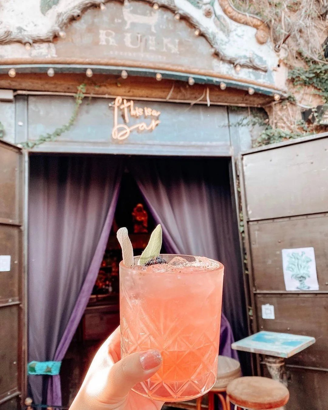 A cocktail held up in front of the curtained doorway of Ruin Bar, with the RUIN sign above.
