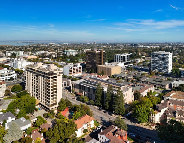 Aerial view of a California city with mid-rise buildings, tree-lined streets, and a distant coastline.