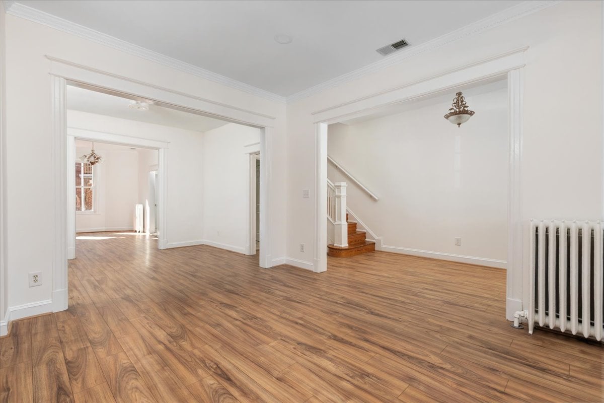Empty room with warm wood floors, wide openings, a staircase, and a white radiator.
