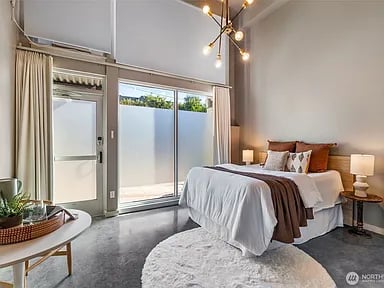 Modern bedroom with a white bed, floor-to-ceiling patio doors, concrete floors, and a brass chandelier.