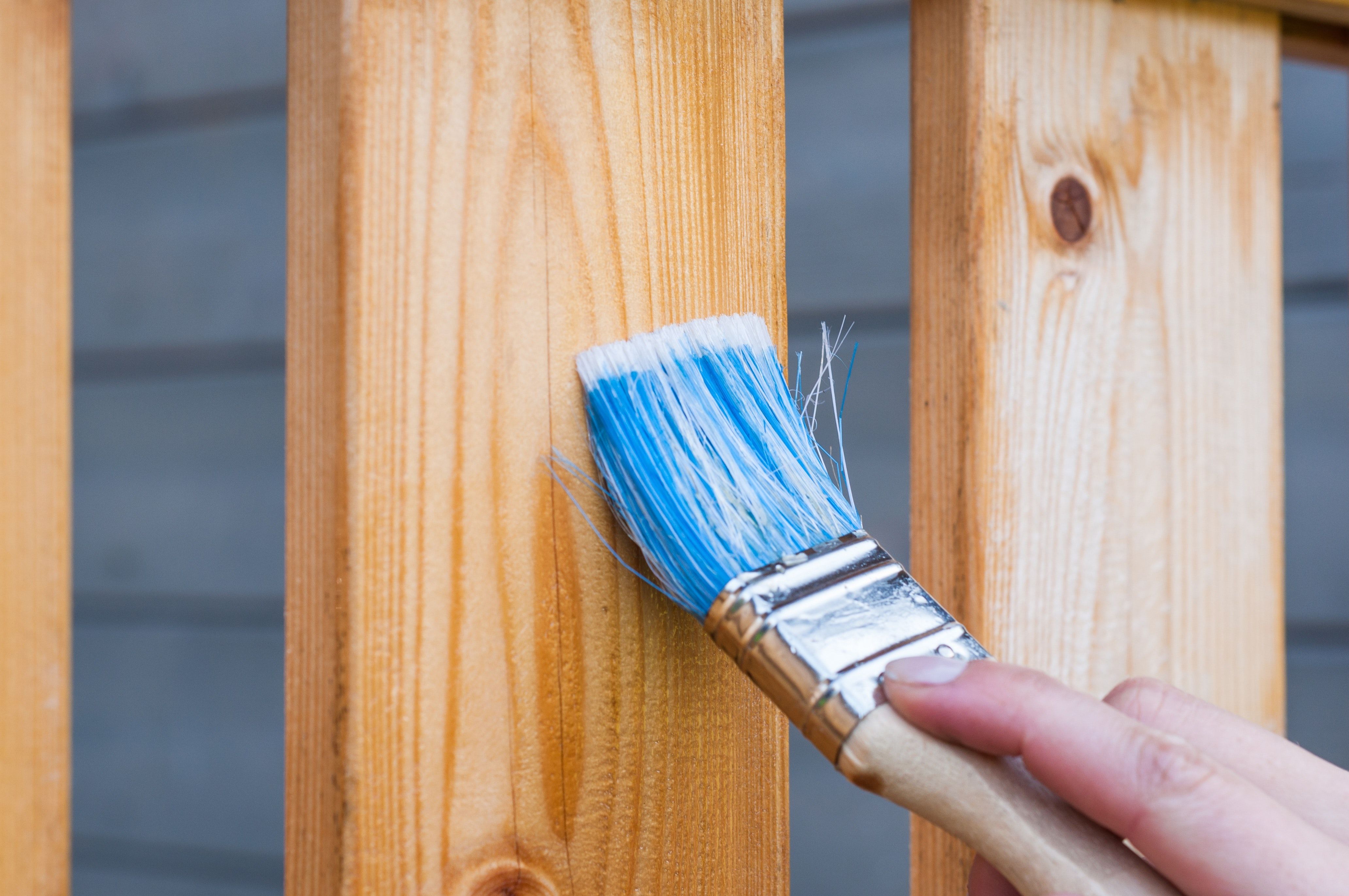 A hand uses a blue-bristled paintbrush on a wooden fence.