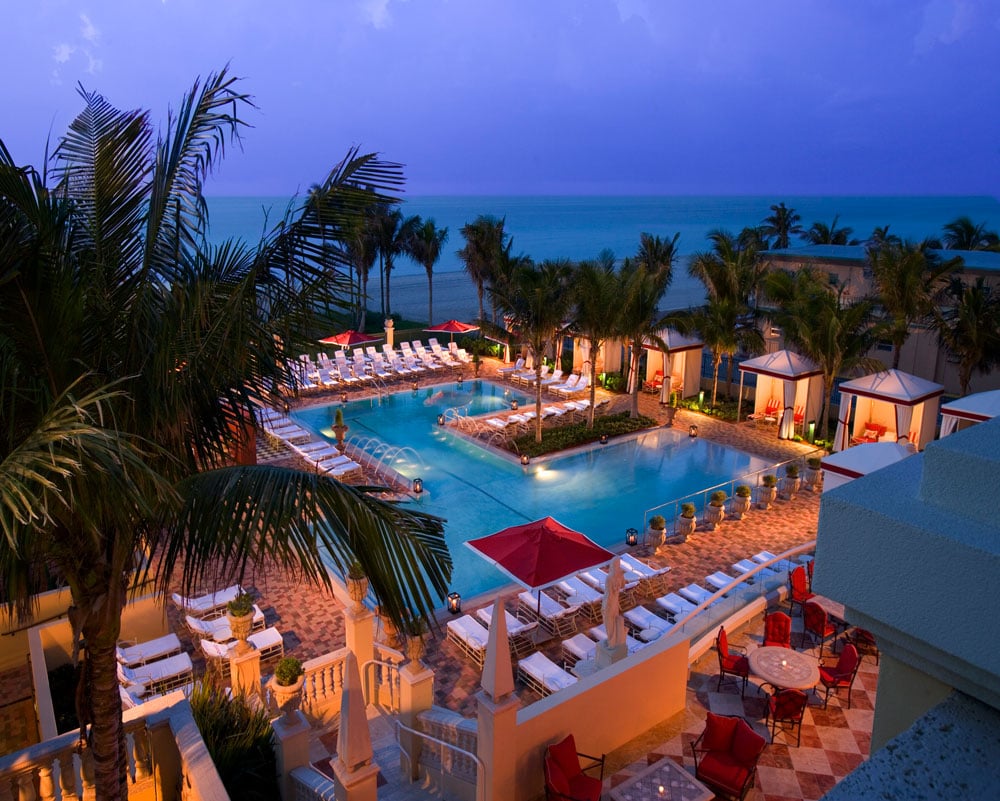 Beachfront resort pool deck at dusk, with palm trees, cabanas, and ocean views.