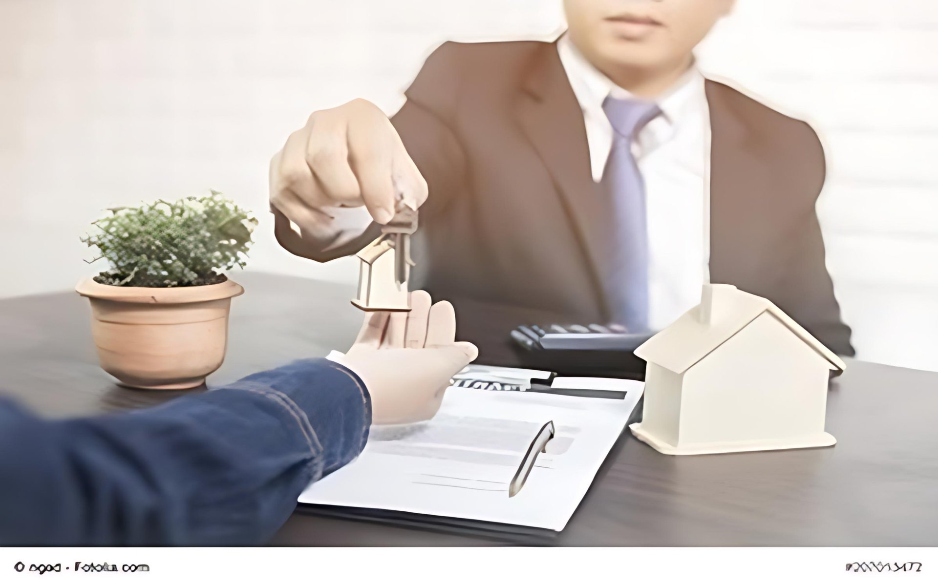 A real estate agent hands keys to a client across a desk beside a model house and paperwork.