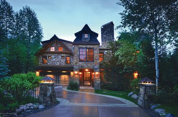 Stone house with illuminated windows, a curved driveway, and stone gate pillars at dusk.