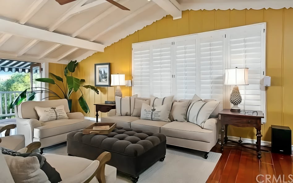 Living room with cream sofas, a dark tufted ottoman, yellow walls, and white plantation shutters.