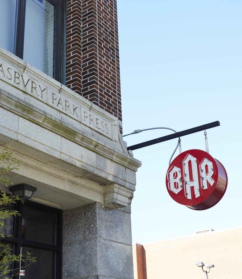 Brick and stone building facade with carved Asbury Park Press lettering and a projecting red BAR sign.