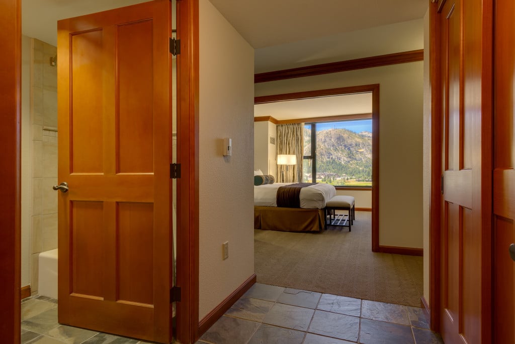 Furnished bedroom suite with a large window overlooking mountains, seen through an open wooden doorway.