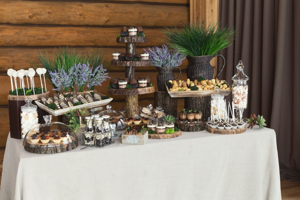 Dessert buffet on a white tablecloth with rustic wooden stands, greenery, and purple flowers.