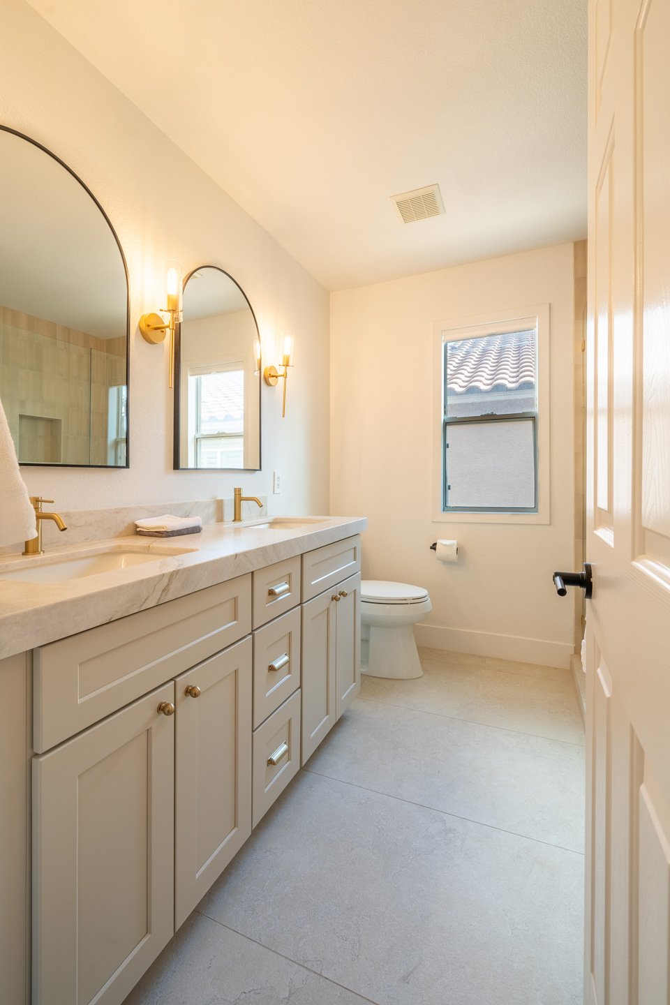 Bathroom with a double vanity, marble-look counter, arched mirrors, and gold-toned fixtures.