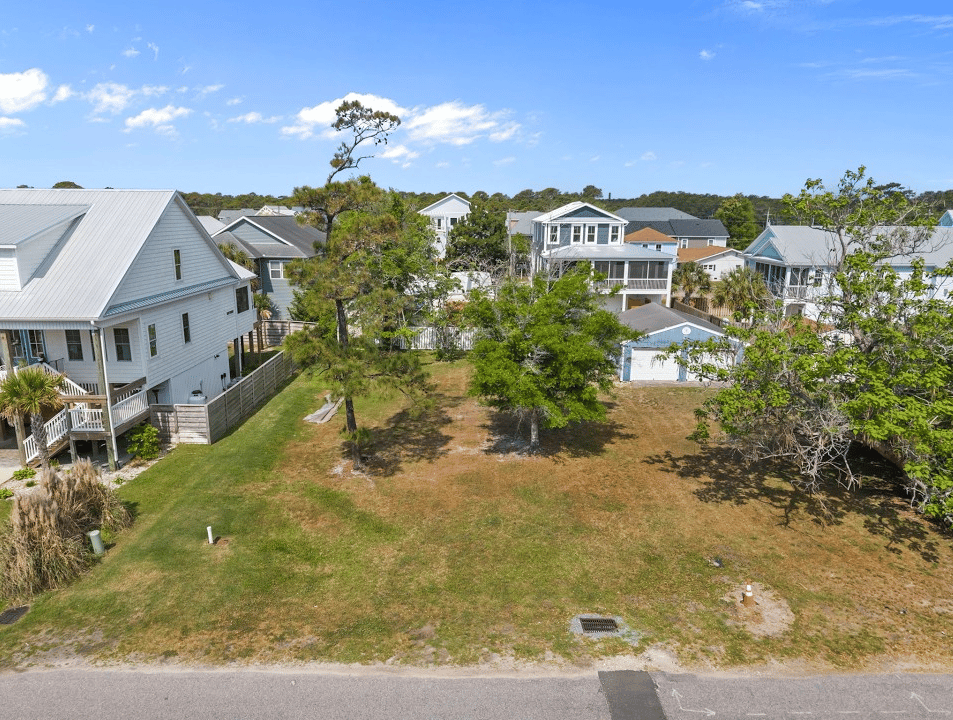 Aerial view of a grassy vacant lot between coastal homes, with the street along its front edge.