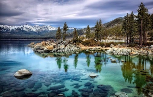 Turquoise mountain lake with rocky shores, pine trees, and snow-covered peaks.