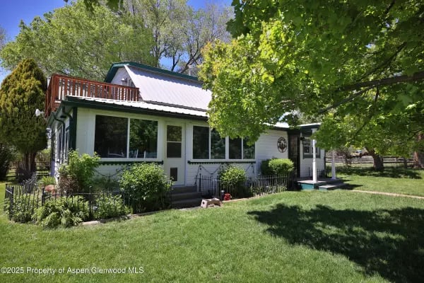 Light-colored house with a metal roof, wooden deck, and tree-shaded lawn.