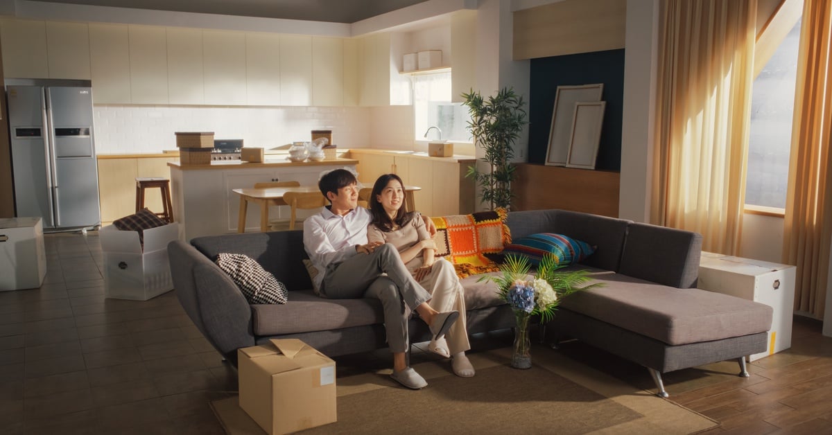 A young couple together on a couch in the living room of a mostly empty home with boxes of unpacked things around them.