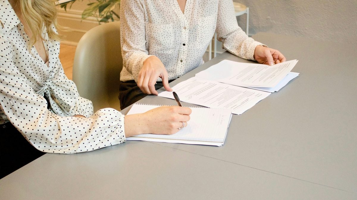 Two women review paperwork at a table as one writes in a notebook.